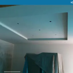 Wnętrze w trakcie wykańczania: sufit z podwieszaną konstrukcją i punktowym oświetleniem, ściany częściowo przygotowane do malowania, meble zabezpieczone folią.