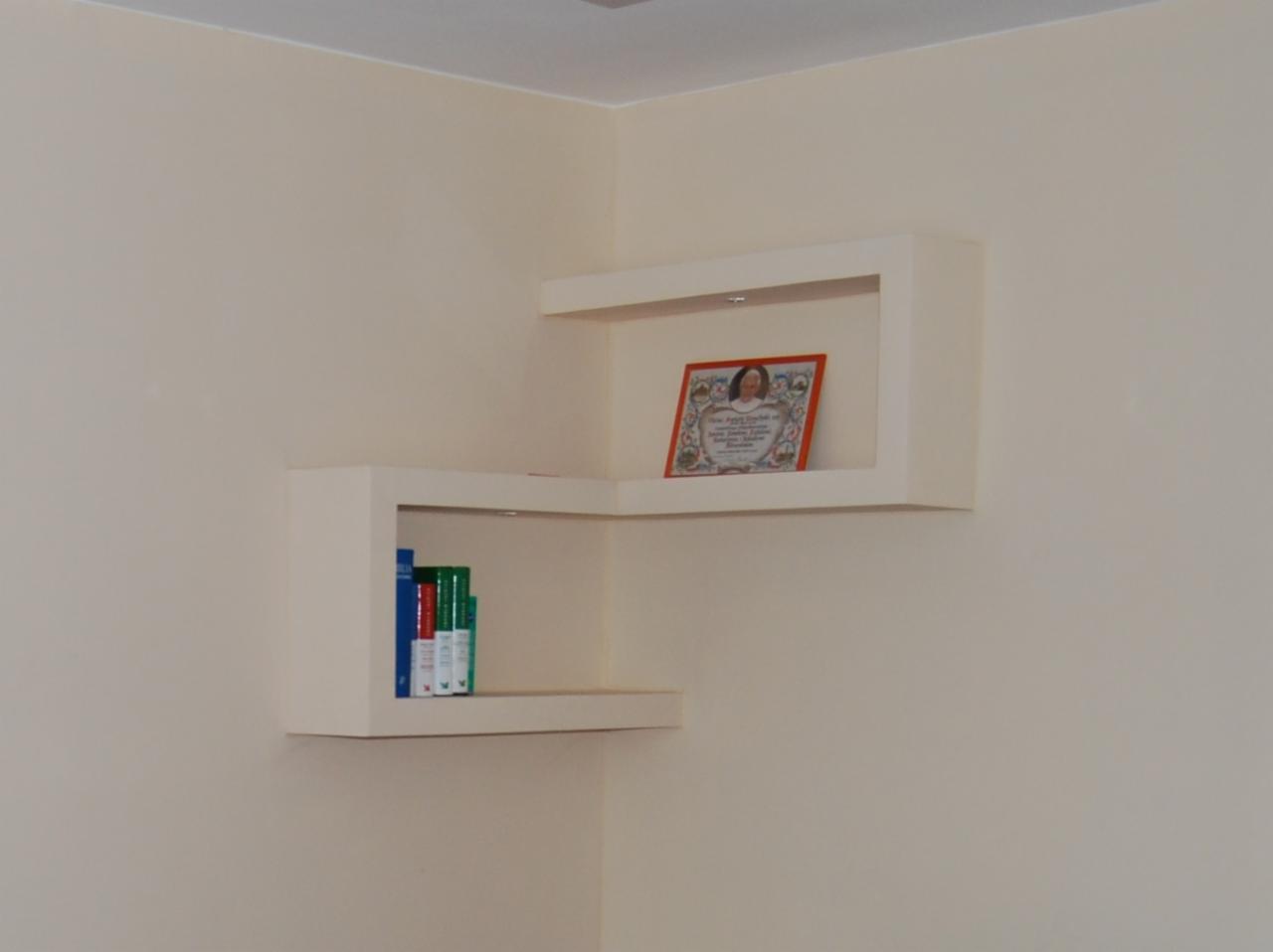 Białe, narożne półki z płyty karton gips, podświetlane od góry, na ścianie w jasnym odcieniu beżu, na jednej półce książki, na drugiej oprawiony obrazek.