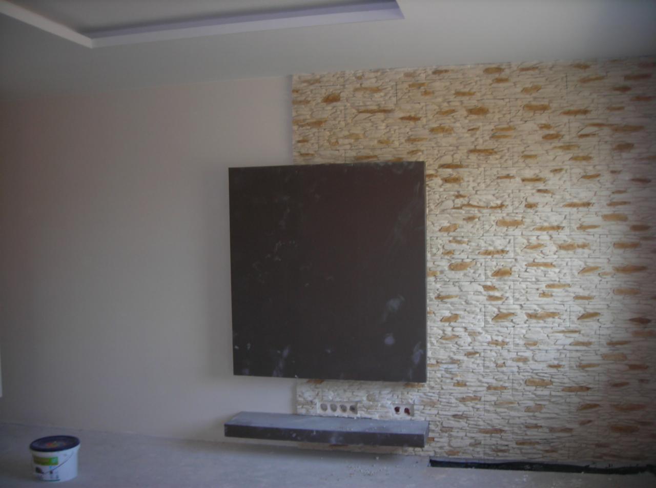 Fragment pokoju z dekoracyjną ścianą imitującą kamień, centralnie umieszczony ciemny panel i półka, widoczny pojemnik z farbą w rogu.