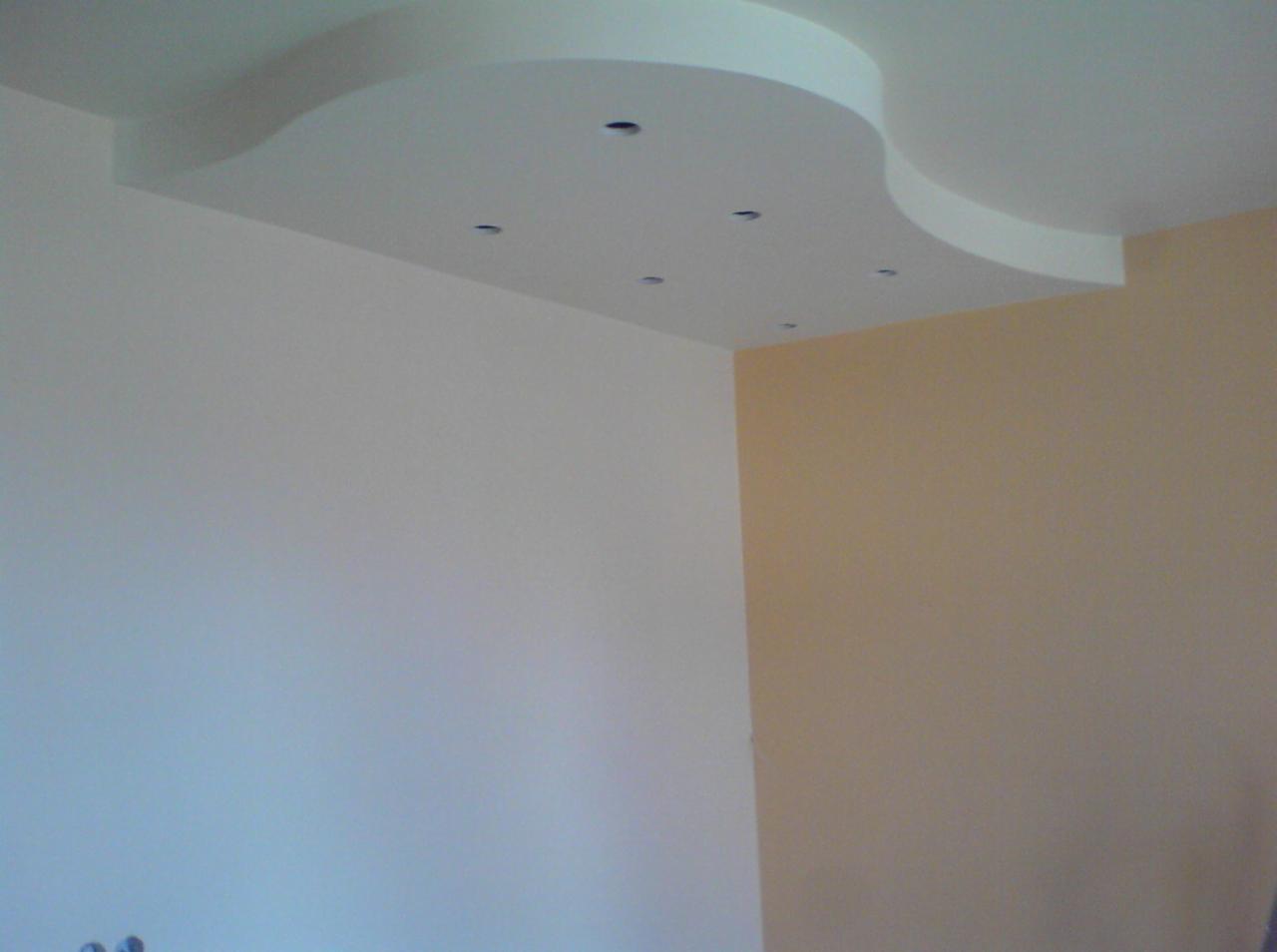 Biały sufit podwieszany o nieregularnym kształcie z wbudowanymi punktowymi oprawami oświetleniowymi, widok na pomalowane ściany w odcieniach bieli i beżu.