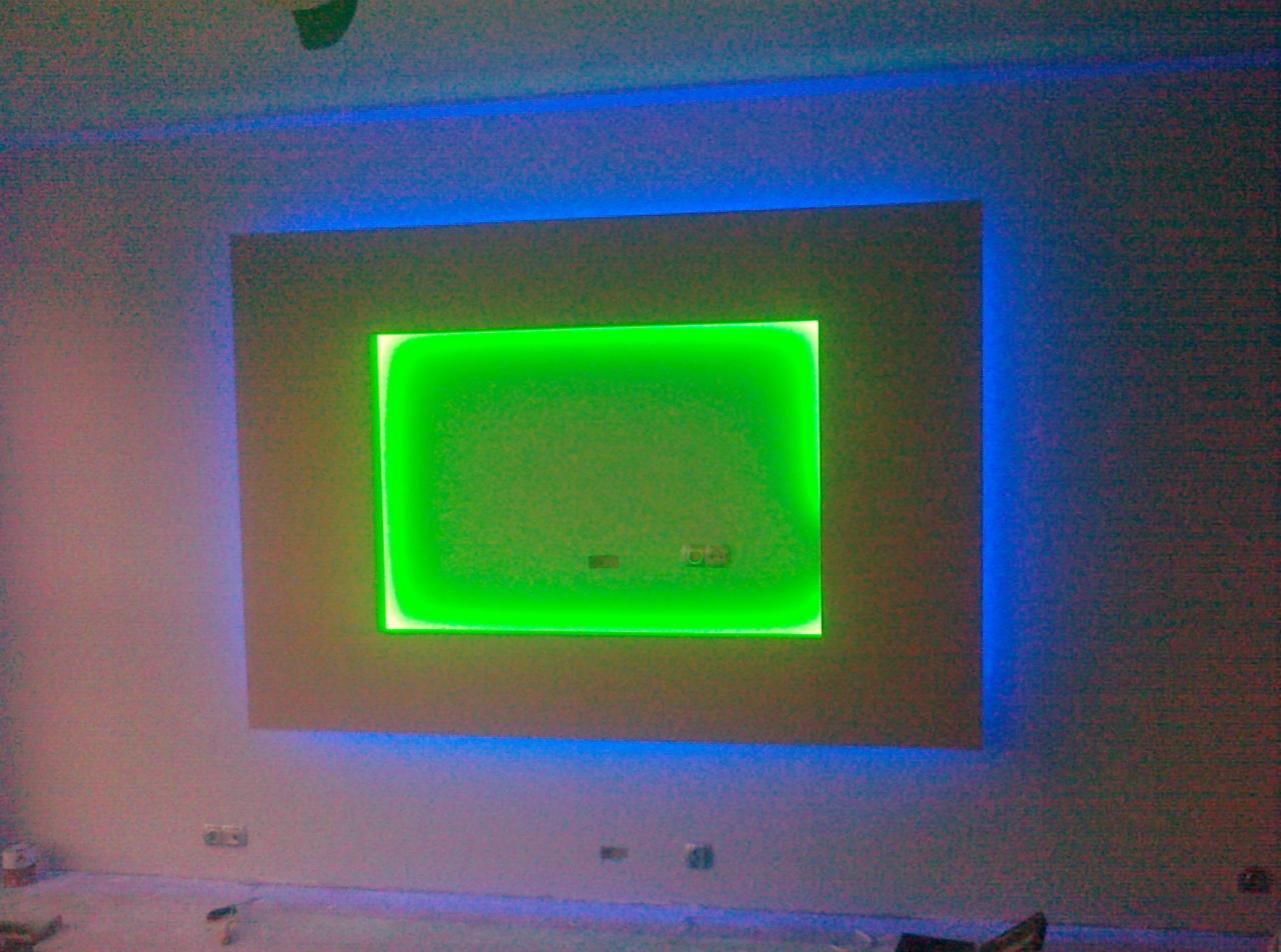 Podświetlany panel dekoracyjny na ścianie z niebieskim i zielonym oświetleniem LED, widoczne gniazdka elektryczne w ścianie, pomieszczenie w trakcie prac wykończeniowych.