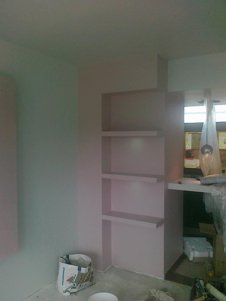 Wnęka z półkami w trakcie tworzenia, ściany pomalowane na jasny róż, widoczny worek po zaprawie i narzędzia malarskie.