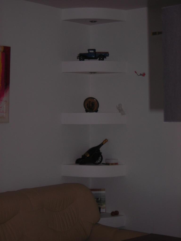 Narożne półki z płyty gipsowo-kartonowej, na których stoją dekoracyjne przedmioty: model samochodu, talerz, figurka anioła i butelka wina. Widoczny fragment sofy.