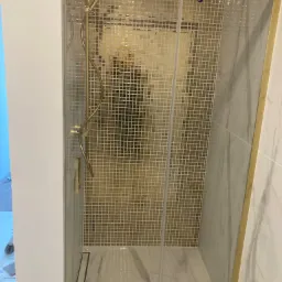 Kabina prysznicowa z drobną mozaiką w odcieniach złota, złote wykończenia, odpływ liniowy, widoczne fragmenty prac wykończeniowych na ścianie.
