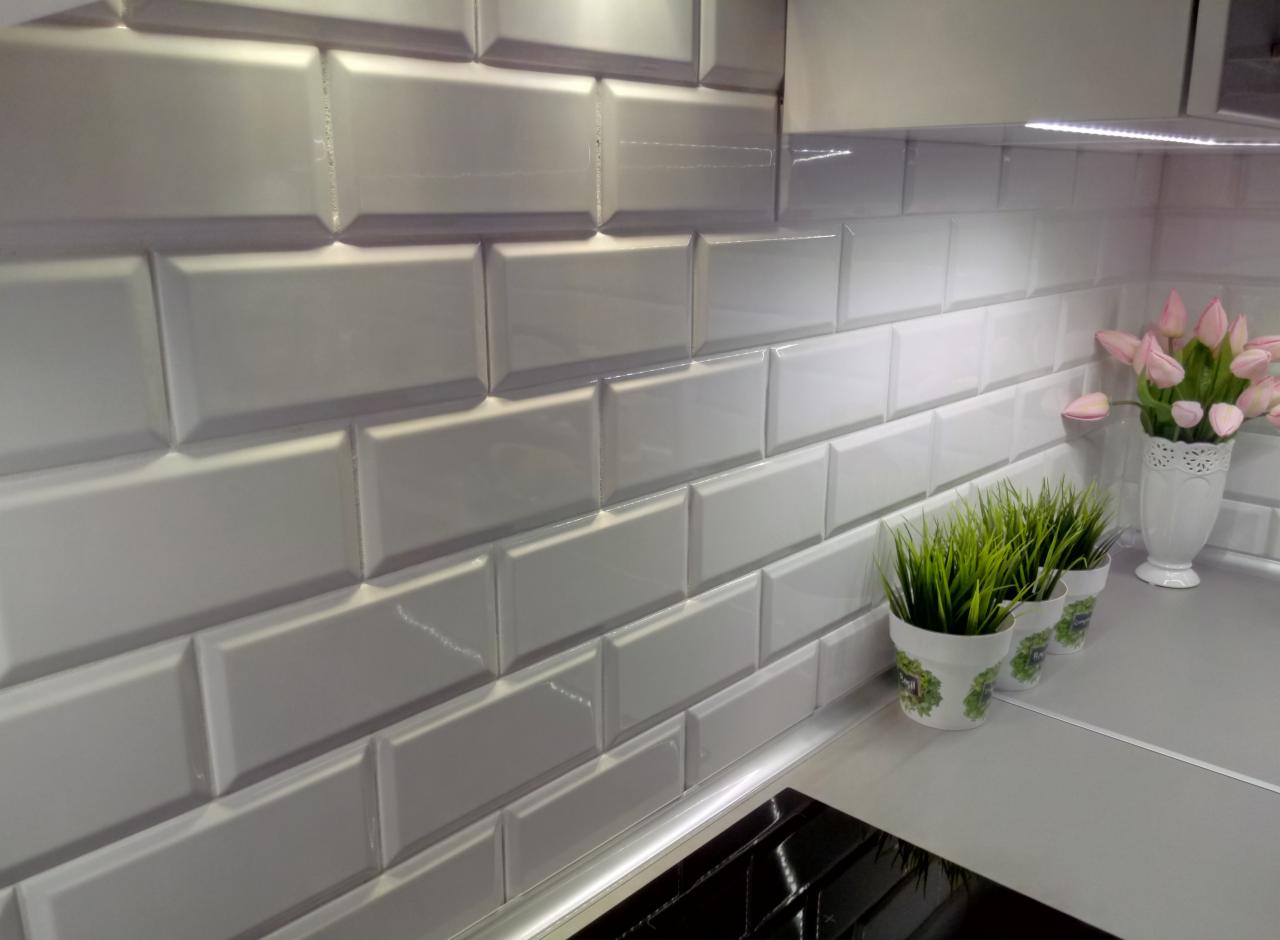 Białe, błyszczące płytki ułożone w cegiełkę na ścianie w kuchni, z trzema doniczkami z zieloną trawą i wazonem z różowymi tulipanami na blacie