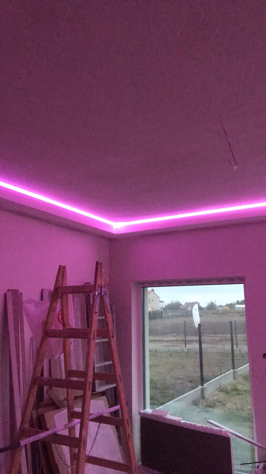Pokój w trakcie prac wykończeniowych z zamontowanym oświetleniem LED w kolorze różowym, drabina oparta o ścianę, widok na zewnątrz przez okno.