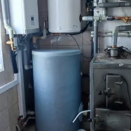 Kompaktowa kotłownia z dwoma białymi zasobnikami ciepłej wody, szarym zbiornikiem akumulacyjnym i starym piecem na paliwo stałe, widoczne liczne rury i zawory.