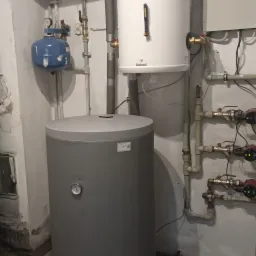 Szary i biały zasobnik ciepłej wody użytkowej Galmet w piwnicy, widoczne rury instalacji, manometr, zawory i sterownik pompy obiegowej.