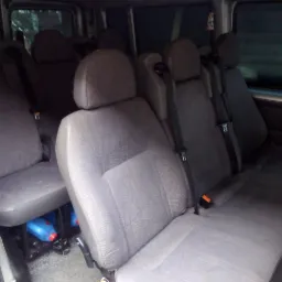 Wnętrze busa z rzędami szarych foteli z zagłówkami i pasami bezpieczeństwa; pod siedzeniami widoczne niebieskie pojemniki z płynem.