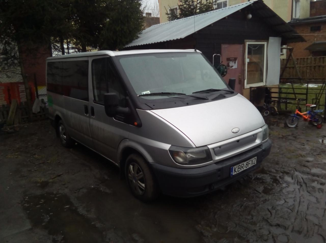Srebrny Ford Transit na błotnistym podwórku z widocznym numerem rejestracyjnym KBR 8FX2, w tle drewniana szopa i dziecięcy rowerek.