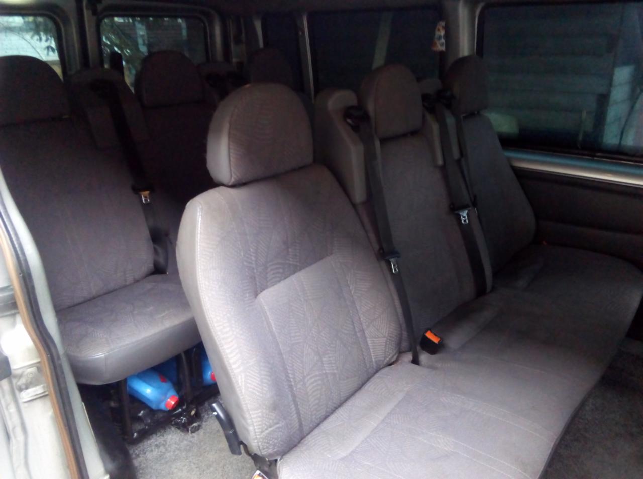 Wnętrze busa z rzędami szarych foteli z zagłówkami i pasami bezpieczeństwa; pod siedzeniami widoczne niebieskie pojemniki z płynem.