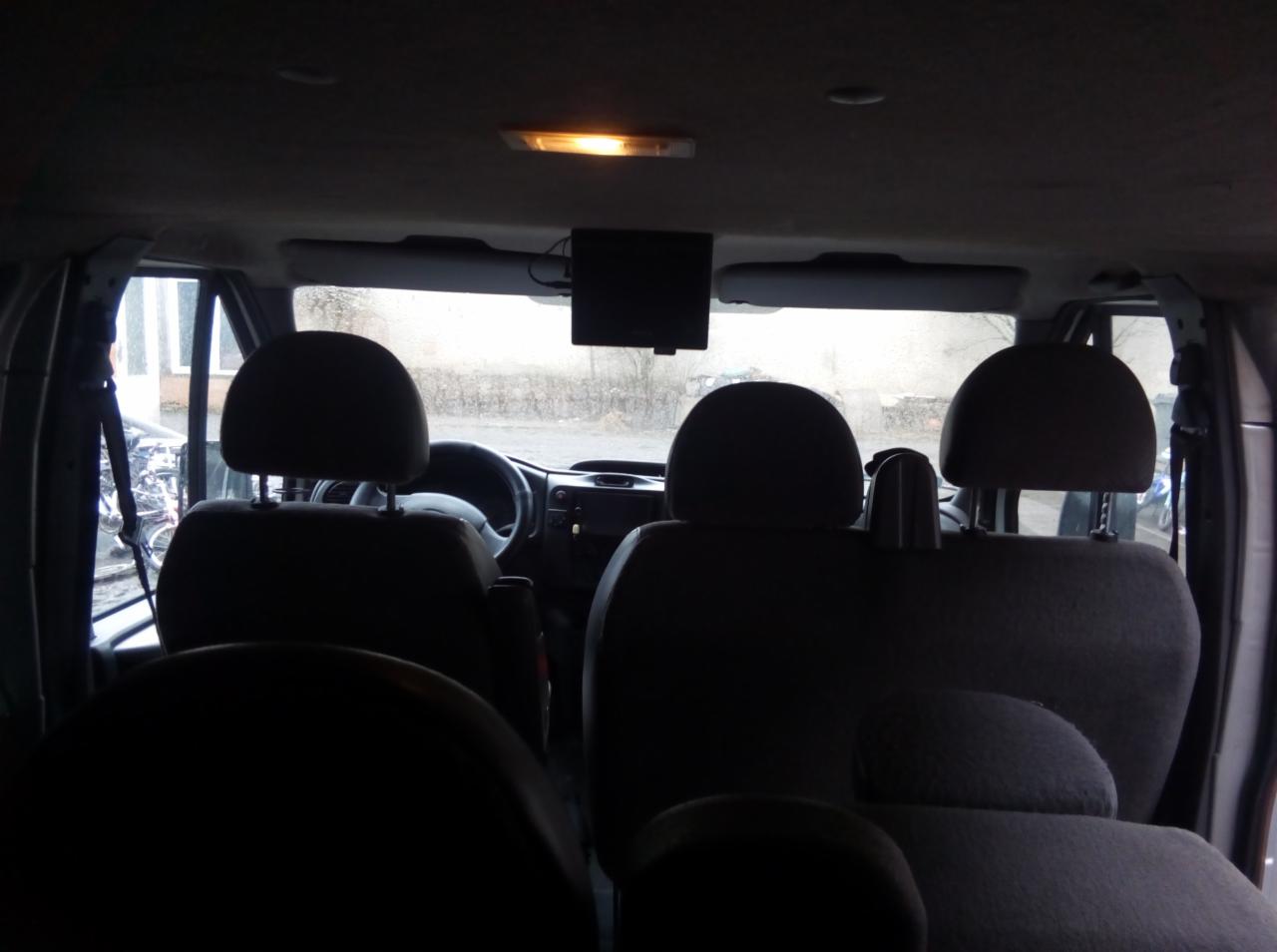 Wnętrze busa z widocznymi fotelami pasażerów, zamontowanym ekranem LCD i elementami kokpitu kierowcy w tle.