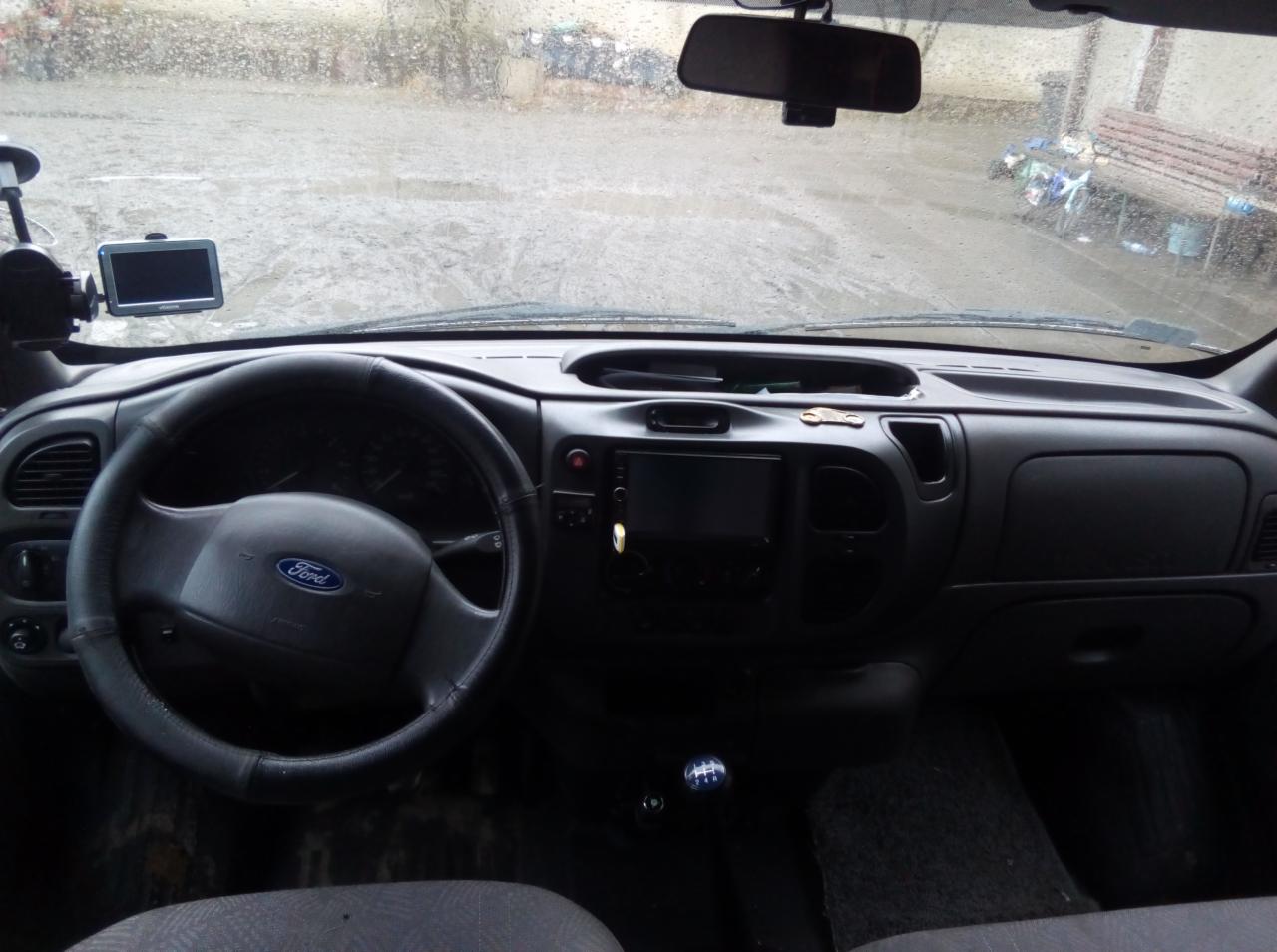 Wnętrze samochodu Ford Transit z widocznym kokpitem, nawigacją GPS i dźwignią zmiany biegów na tle deszczowej pogody za szybą.