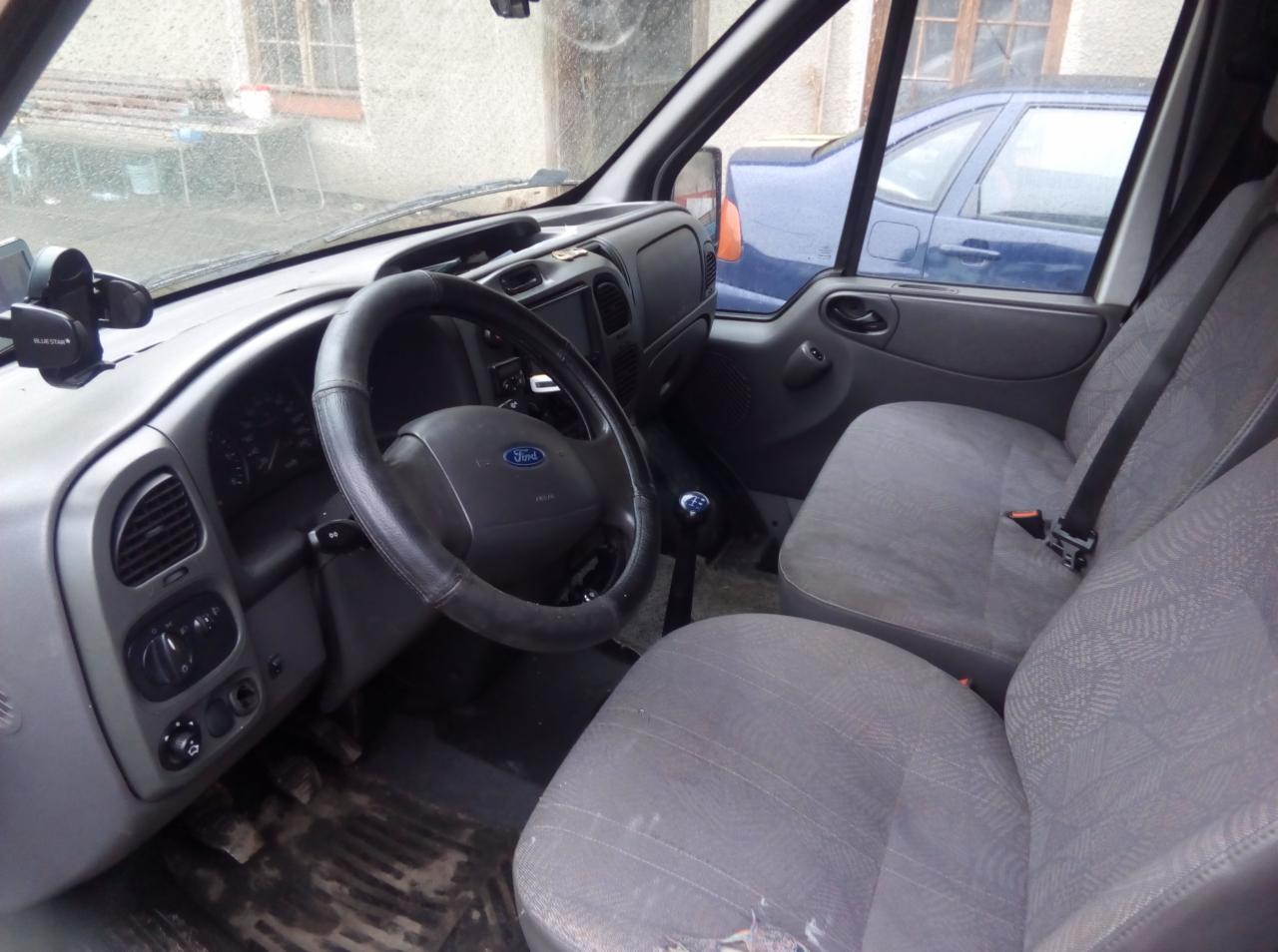 Wnętrze busa Ford Transit z widocznymi szarymi fotelami, deską rozdzielczą i kierownicą z logo Forda. Na przedniej szybie uchwyt na telefon. Za oknem widoczny inny samochód.