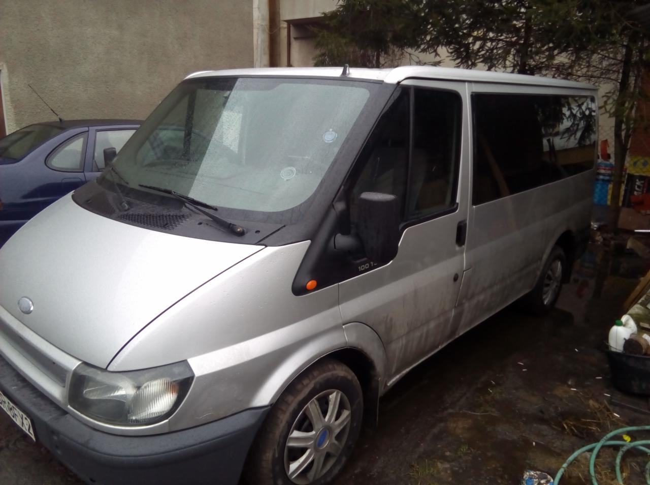 Srebrny bus Ford Transit 100 T z widocznymi śladami błota na karoserii, zaparkowany na zewnątrz budynku.