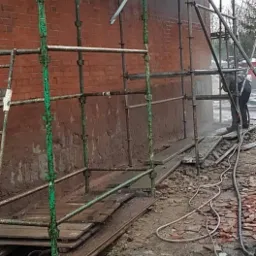 Pracownik w kombinezonie czyszczący ceglaną ścianę budynku pod rusztowaniem, strumień wody pod ciśnieniem usuwa zabrudzenia, widoczny fragment elewacji po czyszczeniu.