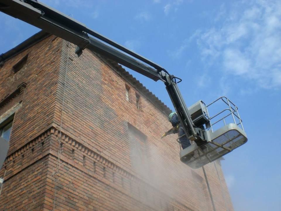 Pracownik w podnośniku koszowym czyści ceglaną elewację budynku strumieniem pod ciśnieniem, z widocznymi śladami czyszczenia i fragmentem błękitnego nieba w tle.