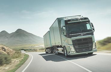 Zielony ciągnik siodłowy Volvo z naczepą jadący po krętej asfaltowej drodze z widokiem na góry w tle, dynamiczne ujęcie z niskiej perspektywy.