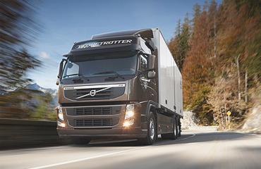 Brązowy samochód ciężarowy Volvo FH z białą naczepą na zakręcie asfaltowej drogi, widok z dołu, w tle las i góry pod błękitnym niebem.