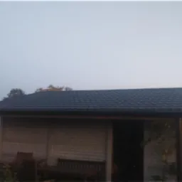 Mała, zadaszona konstrukcja z ciemną, modułową dachówką, widoczna w ogrodzie obok szklarni i betoniarki.