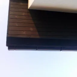 Fragment dachu z ciemnego drewna z widocznymi rynnami w kolorze grafitowym, sfotografowany z dołu na tle jasnego nieba.