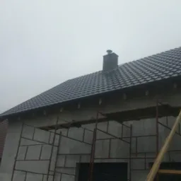 Dom w trakcie krycia dachu blachodachówką w kolorze grafitowym, widoczne rusztowania i komin, pochmurne niebo w tle.