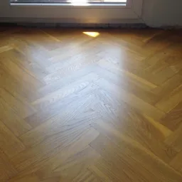 Ułożony parkiet w jodełkę, widoczny fragment okna i ściana z ubytkami w tynku.