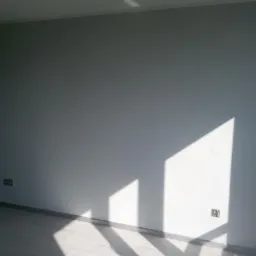 Świeżo pomalowana, jasnoszara ściana z widocznymi promieniami słonecznymi tworzącymi geometryczny wzór, gładka powierzchnia, widoczne gniazdka elektryczne, fragment podłogi z panelami i drzwi szafy...