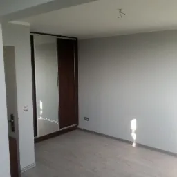 Wnętrze pokoju po remoncie z szarą, gładką ścianą, drewnianą podłogą i szafą wnękową z przesuwanymi drzwiami z lustrem i ciemnego drewna.