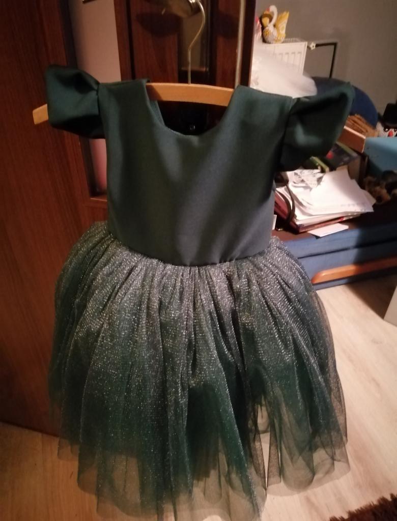 Ciemnozielona sukienka dziewczęca z tiulowym dołem, wisząca na drewnianym wieszaku w pokoju.