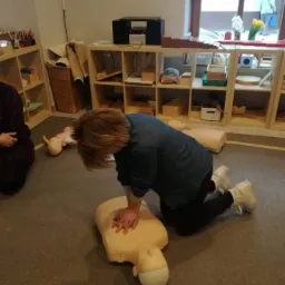 Instruktorka demonstruje resuscytację krążeniowo-oddechową na manekinie podczas zajęć z pierwszej pomocy; widoczne dłonie uciskające klatkę piersiową manekina, a w tle regały z wyposażeniem sali...