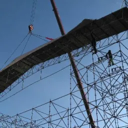 Montaż modułowej konstrukcji rusztowania z użyciem dźwigu, pracownicy na rusztowaniu w kombinezonach, widok z dołu na tle błękitnego nieba i fragmentu budynku.