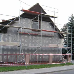 Dom jednorodzinny w trakcie termomodernizacji; widoczne rusztowania, ściana z przyklejonym styropianem i zabezpieczona szpachlą w miejscach kołków mocujących, ogrodzenie posesji z taśmą ostrzegawczą.