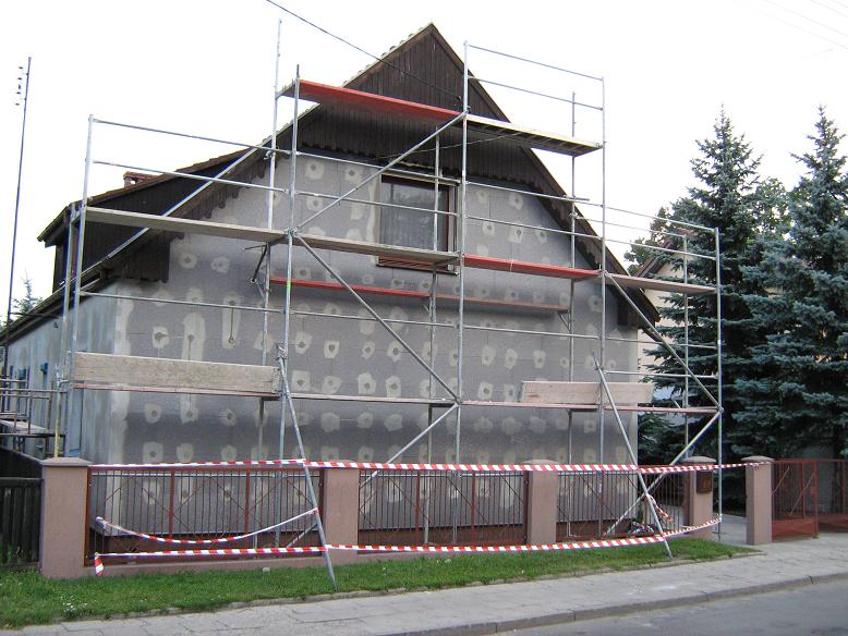 Dom jednorodzinny w trakcie termomodernizacji; widoczne rusztowania, ściana z przyklejonym styropianem i zabezpieczona szpachlą w miejscach kołków mocujących, ogrodzenie posesji z taśmą ostrzegawczą.