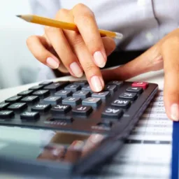 Kobieta z precyzyjnie wypielęgnowanymi dłońmi naciska palcem przycisk na kalkulatorze, obok leży oł&oacute;wek i arkusz kalkulacyjny z danymi finansowymi.