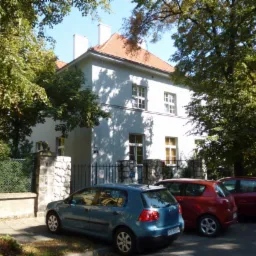 Dwupiętrowy budynek biurowy z białymi ścianami i pomarańczowym dachem, otoczony zielenią drzew, z zaparkowanymi trzema samochodami na pierwszym planie.