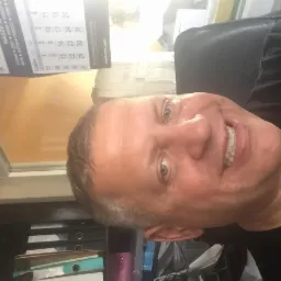 Uśmiechnięty mężczyzna pozujący na tle kalendarza biurowego i regału z dokumentami.