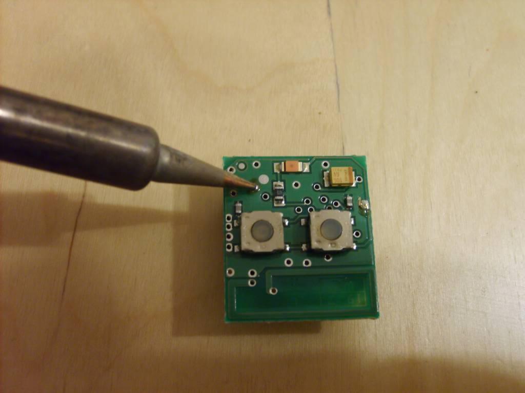 Lutowanie elementów na zielonej płytce drukowanej za pomocą lutownicy, widoczne dwa przyciski i inne komponenty elektroniczne.