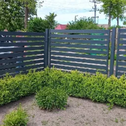 Grafitowe poziome przęsła ogrodzenia z widocznym pojazdem firmy montażowej w tle, obsadzone niskimi, zielonymi krzewami.