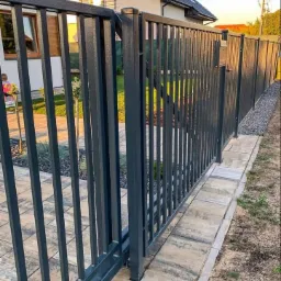 Nowoczesna, przesuwna brama panelowa w kolorze grafitowym, z pionowymi prętami, zamontowana na posesji z kostką brukową i zadbanym trawnikiem.