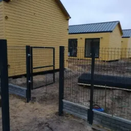 Częściowo zamontowane czarne ogrodzenie panelowe z bramką na tle żółtych domków modułowych, widoczna osoba z łopatą i elementy drenażu.