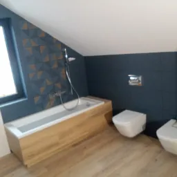 Nowoczesna łazienka na poddaszu z wanną obudowaną drewnem, toaletą i bidetem w stylu minimalistycznym, ciemne płytki na ścianach z geometrycznym wzorem w okolicy okna.