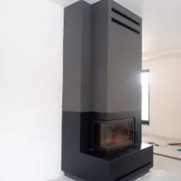 Nowoczesny, dwukolorowy kominek z wkładem kominkowym, z widocznymi kratkami wentylacyjnymi, na tle białej ściany w trakcie prac wykończeniowych.