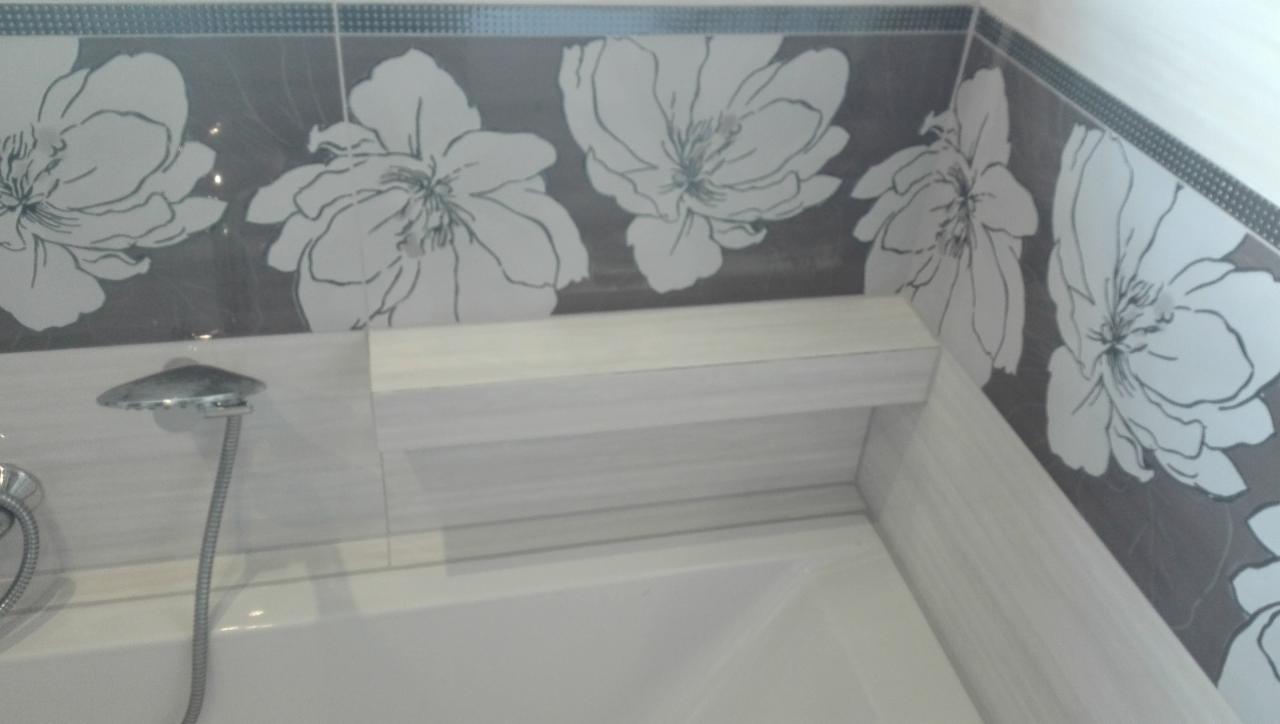 Wykończona łazienka z wanną, półką w zabudowie i szarymi płytkami z motywem kwiatowym, widok na narożnik i słuchawkę prysznicową.