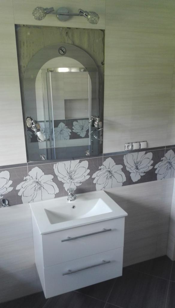 Wykończona łazienka z białą szafką pod umywalkę z dwiema szufladami, lustrem w kształcie łuku z oświetleniem i płytkami z motywem kwiatowym.