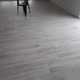 Wykończona podłoga z szarych płytek imitujących drewno, widoczny fragment krzesła i okna.