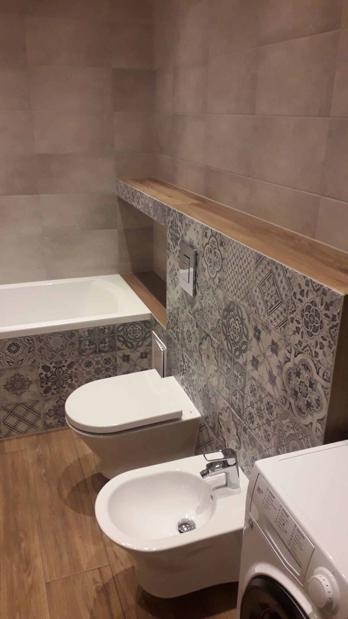 Nowoczesna łazienka z płytkami w stylu patchwork, wiszącą toaletą i bidetem, drewnianą półką nad zabudową oraz pralką w rogu.