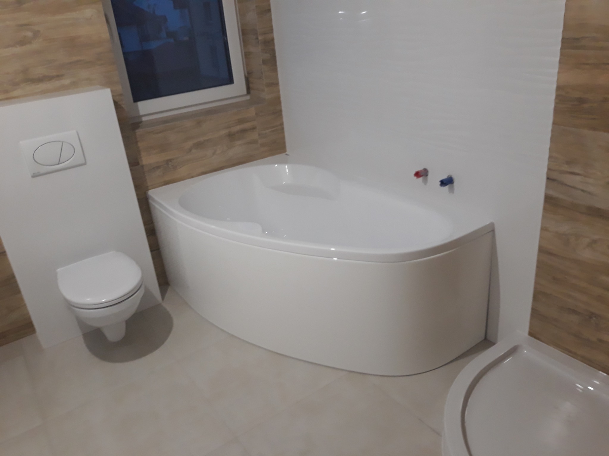 Biała narożna wanna, podwieszana toaleta z panelem spłukującym i częściowo widoczny brodzik w wykończonej łazience z imitacją drewnianych płytek na ścianach.