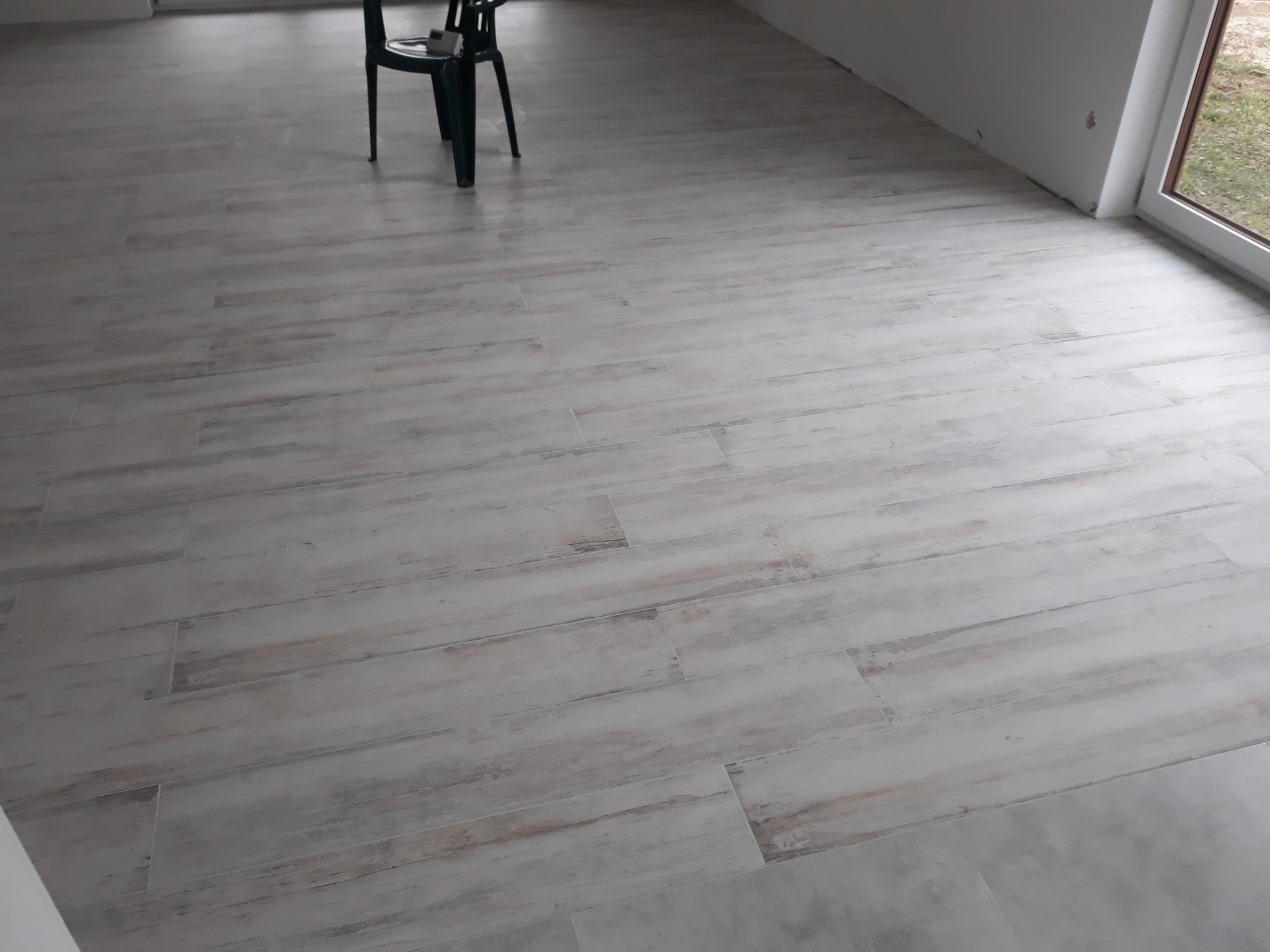 Wykończona podłoga z szarych płytek imitujących drewno, widoczny fragment krzesła i okna.
