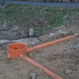 Montaż pomarańczowych rur kanalizacyjnych i zbiornika w wykopie ziemnym, widoczne drzewa w tle.